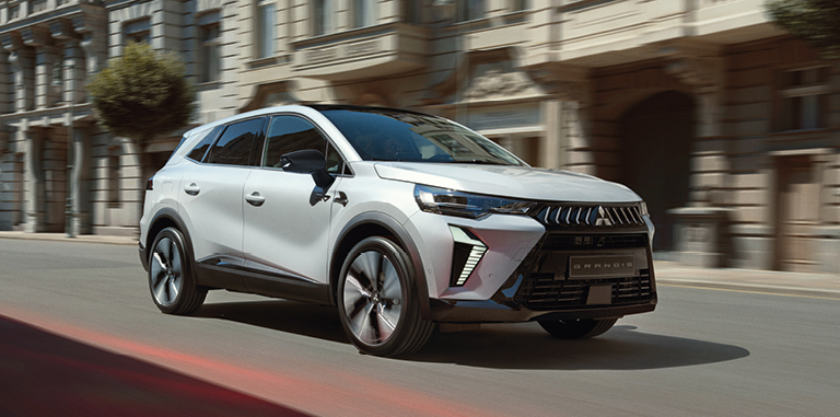Wei&szlig;er Mitsubishi Grandis Kompakt-SUV f&auml;hrt eine Stadtstra&szlig;e entlang, umgeben von historischen Fassaden, leicht verwischt dargestellt, um Bewegung und Fahrdynamik zu betonen.