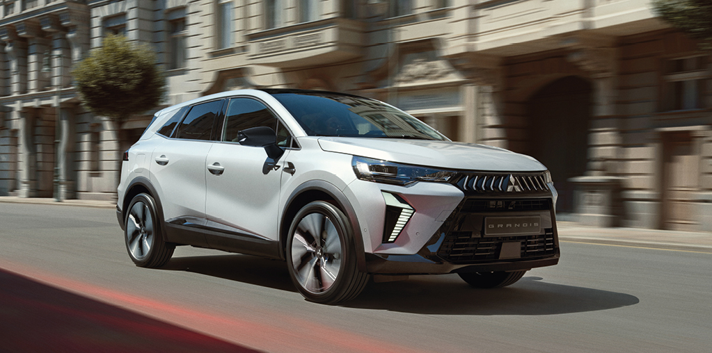 Wei&szlig;er Mitsubishi Grandis Kompakt-SUV f&auml;hrt eine Stadtstra&szlig;e entlang, umgeben von historischen Fassaden, leicht verwischt dargestellt, um Bewegung und Fahrdynamik zu betonen.