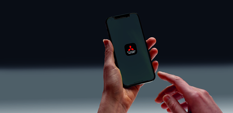 Eine Hand h&auml;lt ein Smartphone vor dunkelgrauem Hintergrund, w&auml;hrend die andere Hand kurz davor ist, den Bildschirm zu ber&uuml;hren. Auf dem Display ist das Icon Mitsubishi Motors App zu sehen.