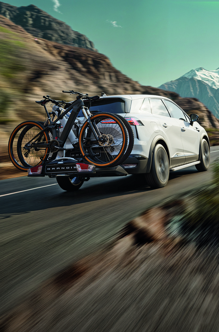 Wei&szlig;er Mitsubishi Grandis f&auml;hrt mit montiertem Heck-Fahrradtr&auml;ger und zwei Fahrr&auml;dern durch eine Berglandschaft auf einer Landstra&szlig;e.