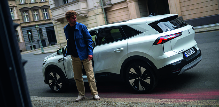 Mitsubishi Grandis Intro Edition – stilvoll ankommen in der Stadt Mann steigt aus einem weißen Mitsubishi Grandis SUV auf einer ruhigen Stadtstraße, Fahrzeug in schräger Rückansicht am Straßenrand geparkt.