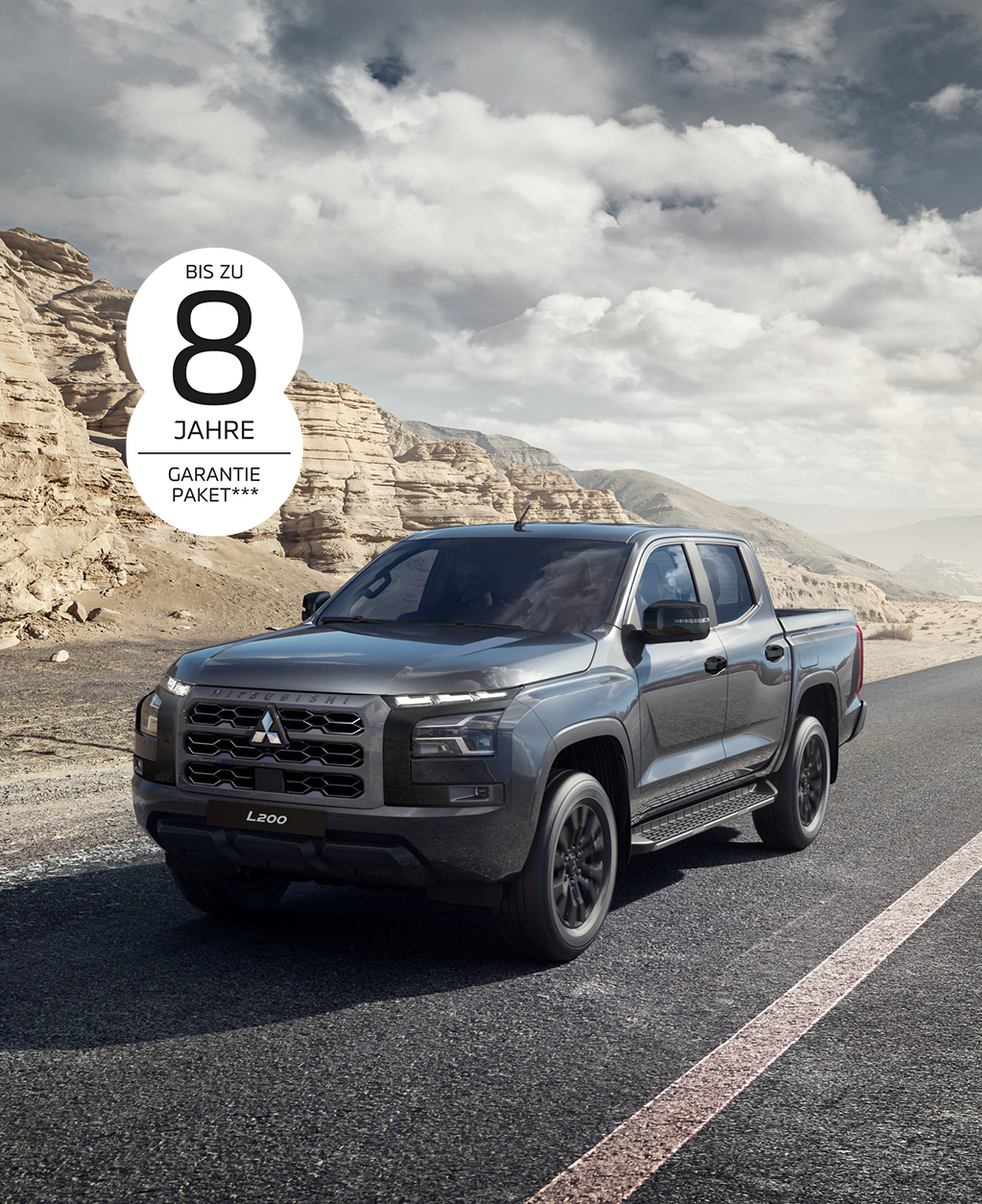 Grauer Mitsubishi L200 Pick-up in Front-Seitenansicht auf einer asphaltierten Stra&szlig;e durch eine karge Felslandschaft; oben links ist der Hinweis &bdquo;Bis zu 8 Jahre Garantiepaket***&ldquo; zu sehen.