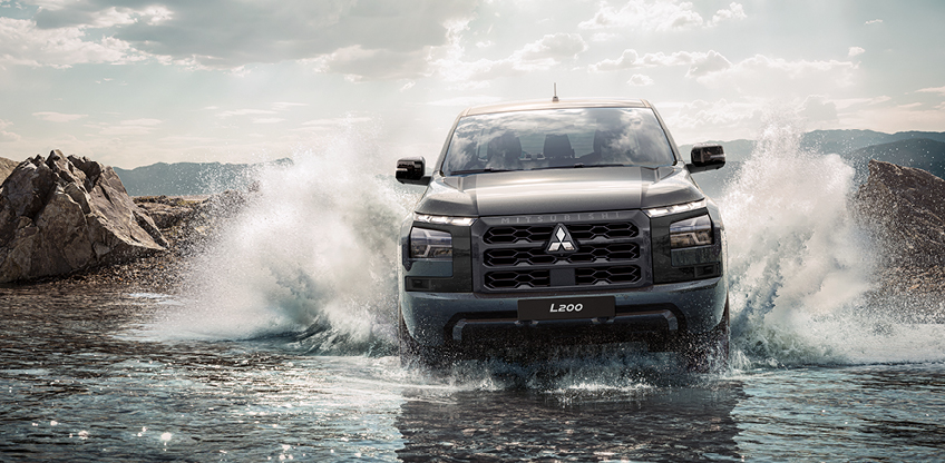 Frontansicht eines schwarzen Mitsubishi L200, der durch seichtes Wasser f&auml;hrt und dabei hohe Spritzfont&auml;nen aufwirbelt. Im Hintergrund sind felsige Ufer und bewaldete Berge zu sehen.