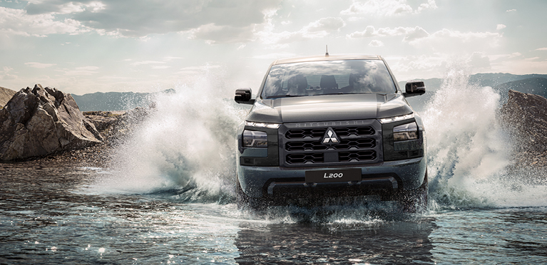 Frontansicht eines schwarzen Mitsubishi L200, der durch seichtes Wasser f&auml;hrt und dabei hohe Spritzfont&auml;nen aufwirbelt. Im Hintergrund sind felsige Ufer und bewaldete Berge zu sehen.