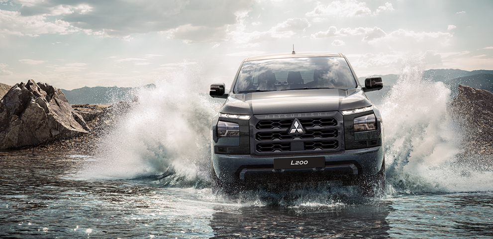 Frontansicht eines schwarzen Mitsubishi L200, der durch seichtes Wasser f&auml;hrt und dabei hohe Spritzfont&auml;nen aufwirbelt. Im Hintergrund sind felsige Ufer und bewaldete Berge zu sehen.