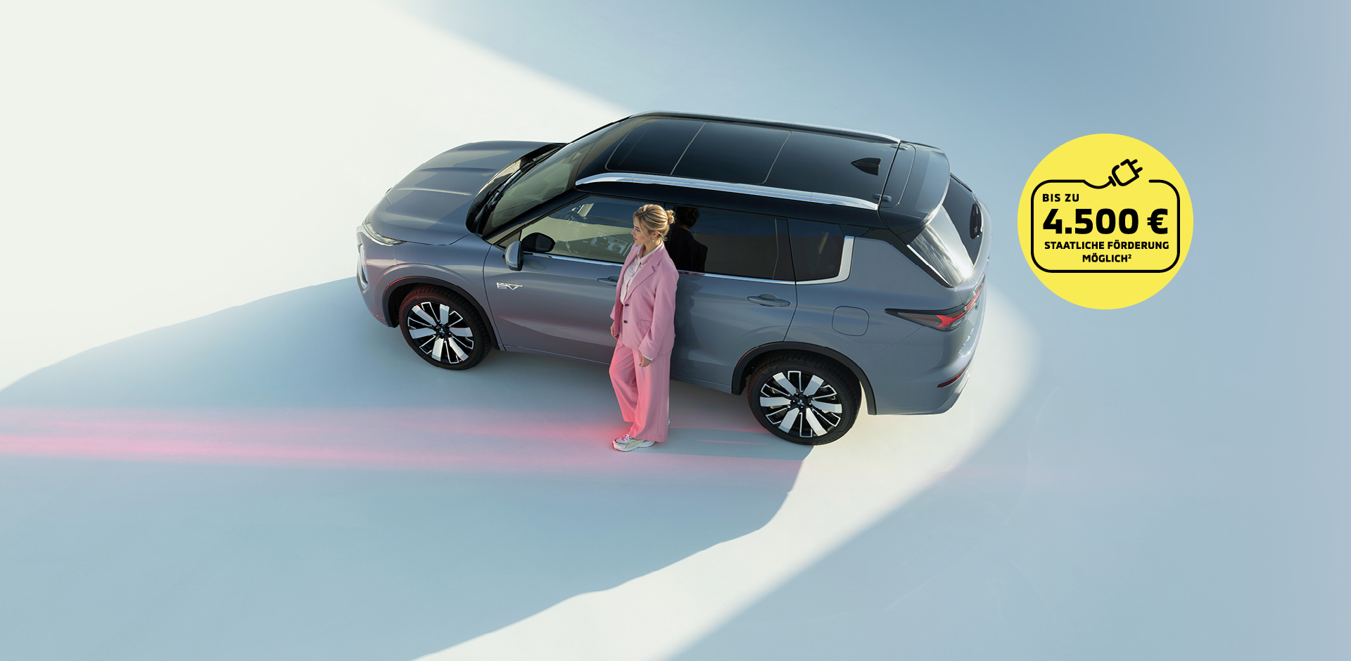 Grauer Mitsubishi Outlander Plug-in Hybrid SUV von oben, Frau in rosafarbenem Anzug steht neben dem Fahrzeug. Gelber Kreis mit Hinweis auf staatliche F&ouml;rderung bis zu 4.500 Euro.