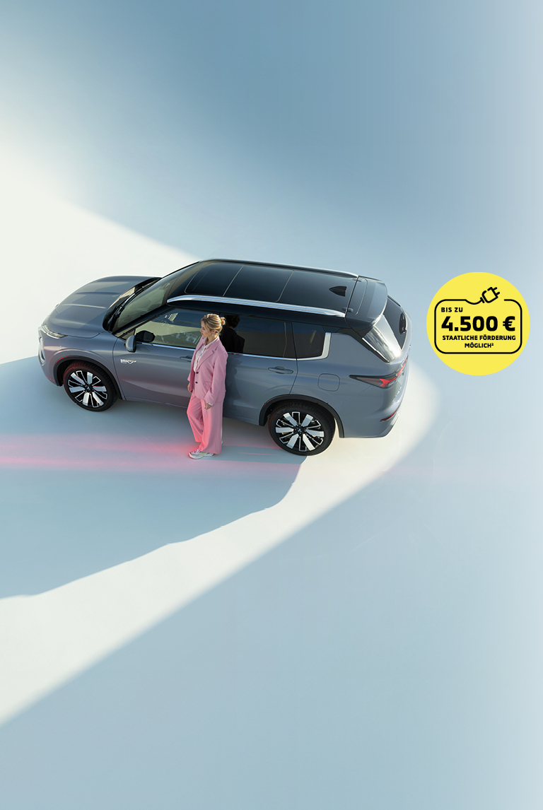 Grauer Mitsubishi Outlander Plug-in Hybrid SUV von oben, Frau in rosafarbenem Anzug steht neben dem Fahrzeug. Gelber Kreis mit Hinweis auf staatliche F&ouml;rderung bis zu 4.500 Euro.