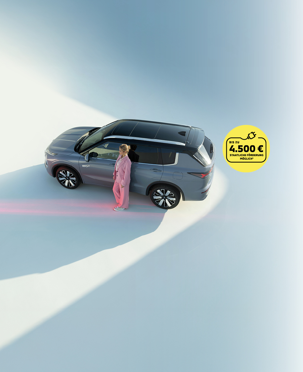 Grauer Mitsubishi Outlander Plug-in Hybrid SUV von oben, Frau in rosafarbenem Anzug steht neben dem Fahrzeug. Gelber Kreis mit Hinweis auf staatliche F&ouml;rderung bis zu 4.500 Euro.