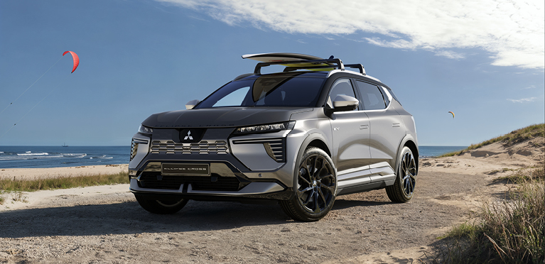 Ein Mitsubishi Eclipse Cross steht auf einem sandigen Weg an der K&uuml;ste, mit einem Surfbrett auf dem Dachtr&auml;ger; im Hintergrund Meer, Strand und bunte Kite-Segel am Himmel.
