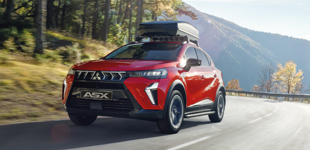Ein roter Mitsubishi Kompakt-SUV mit schwarzem Dach und montierter Dachbox f&auml;hrt bei Tageslicht auf einer kurvigen Bergstra&szlig;e, umgeben von B&auml;umen und H&uuml;geln im Hintergrund.