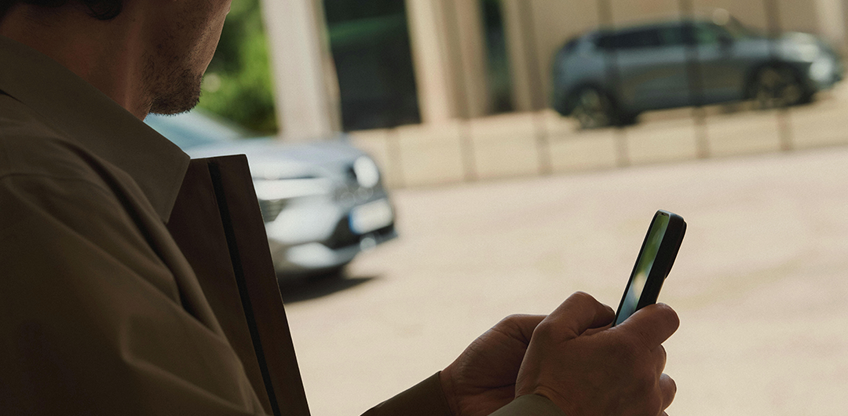 Konnektivität Im Vordergrund hält ein Mann ein Mobiltelefon in der Hand, während sich der Eclipse Cross im Hintergrund in der Glasfront eines modernen Gebäudes spiegelt.