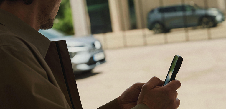 Konnektivität Im Vordergrund hält ein Mann ein Mobiltelefon in der Hand, während sich der Eclipse Cross im Hintergrund in der Glasfront eines modernen Gebäudes spiegelt.