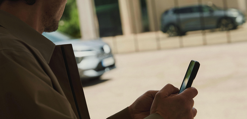 Konnektivität Im Vordergrund hält ein Mann ein Mobiltelefon in der Hand, während sich der Eclipse Cross im Hintergrund in der Glasfront eines modernen Gebäudes spiegelt.